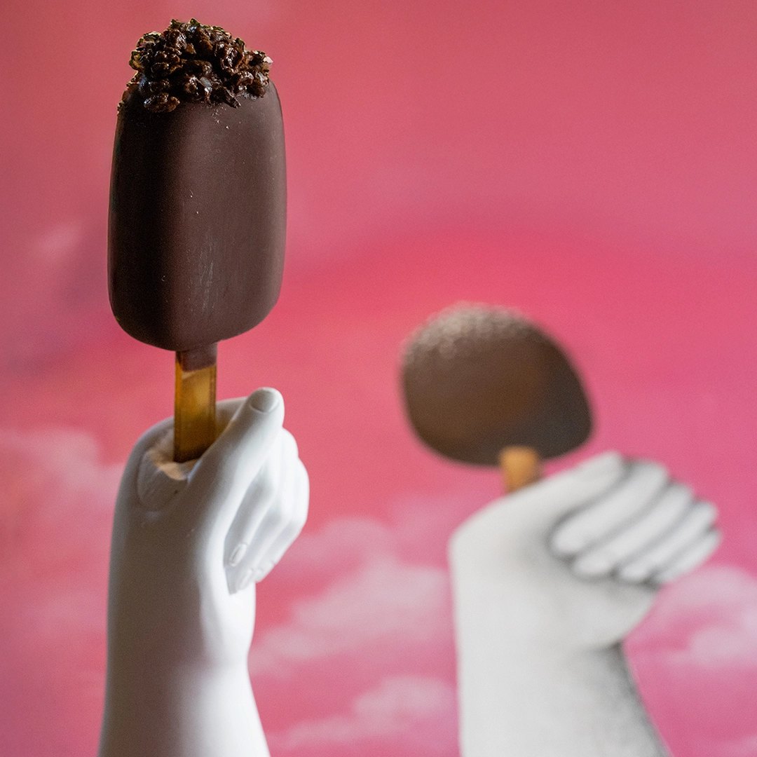 Un dessert a forma di ghiacciolo al cioccolato, tenuto da una mano guantata di bianco su un fondo rosa