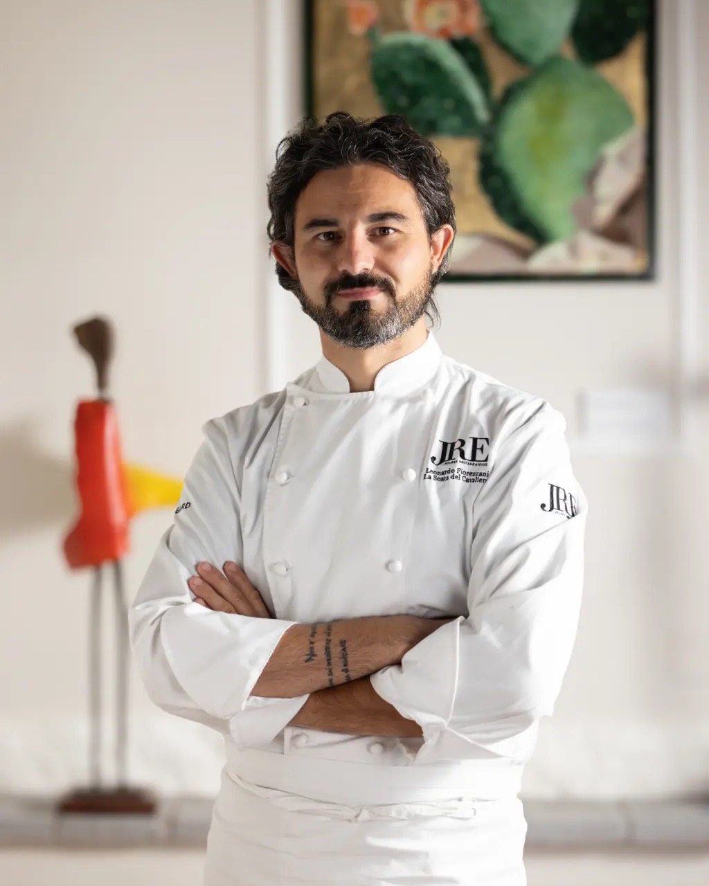 Leonardo Fiorenzani in giacca da cuoco JRE, braccia conserte, davanti a un quadro con fichi d’India